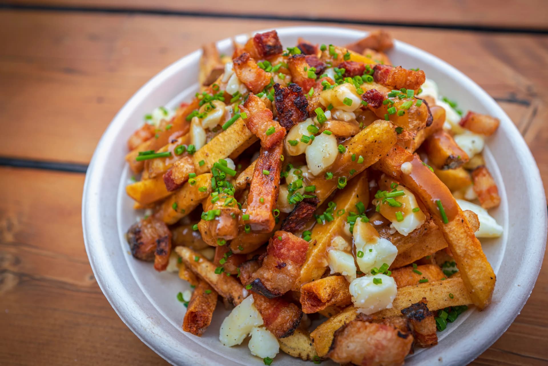 Poutine with fries, cheese curds, gravy, bacon, and chives in a white bowl on a wooden table.