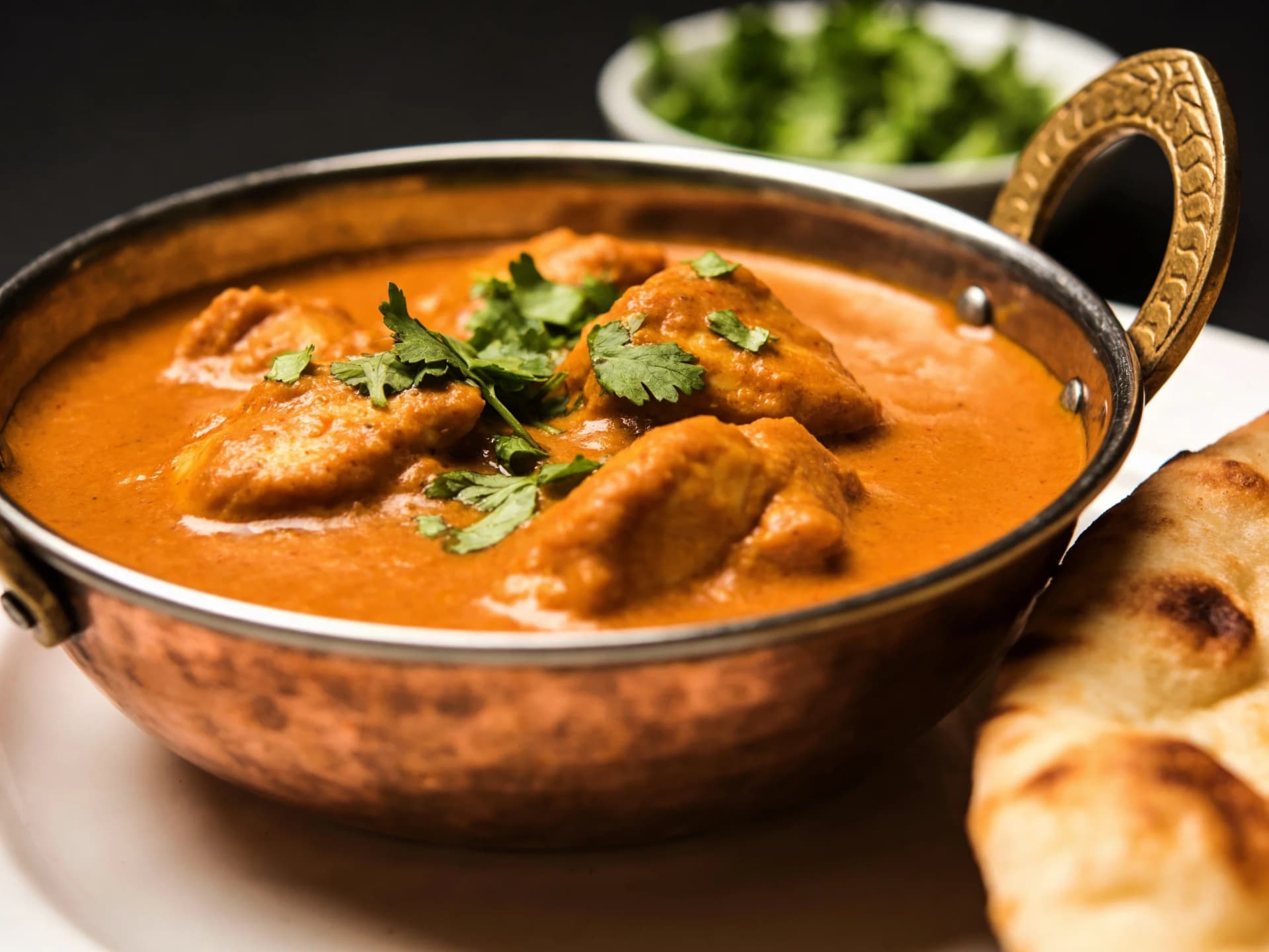 Butter chicken in a copper bowl with creamy orange sauce, garnished with cilantro, served with naan bread.