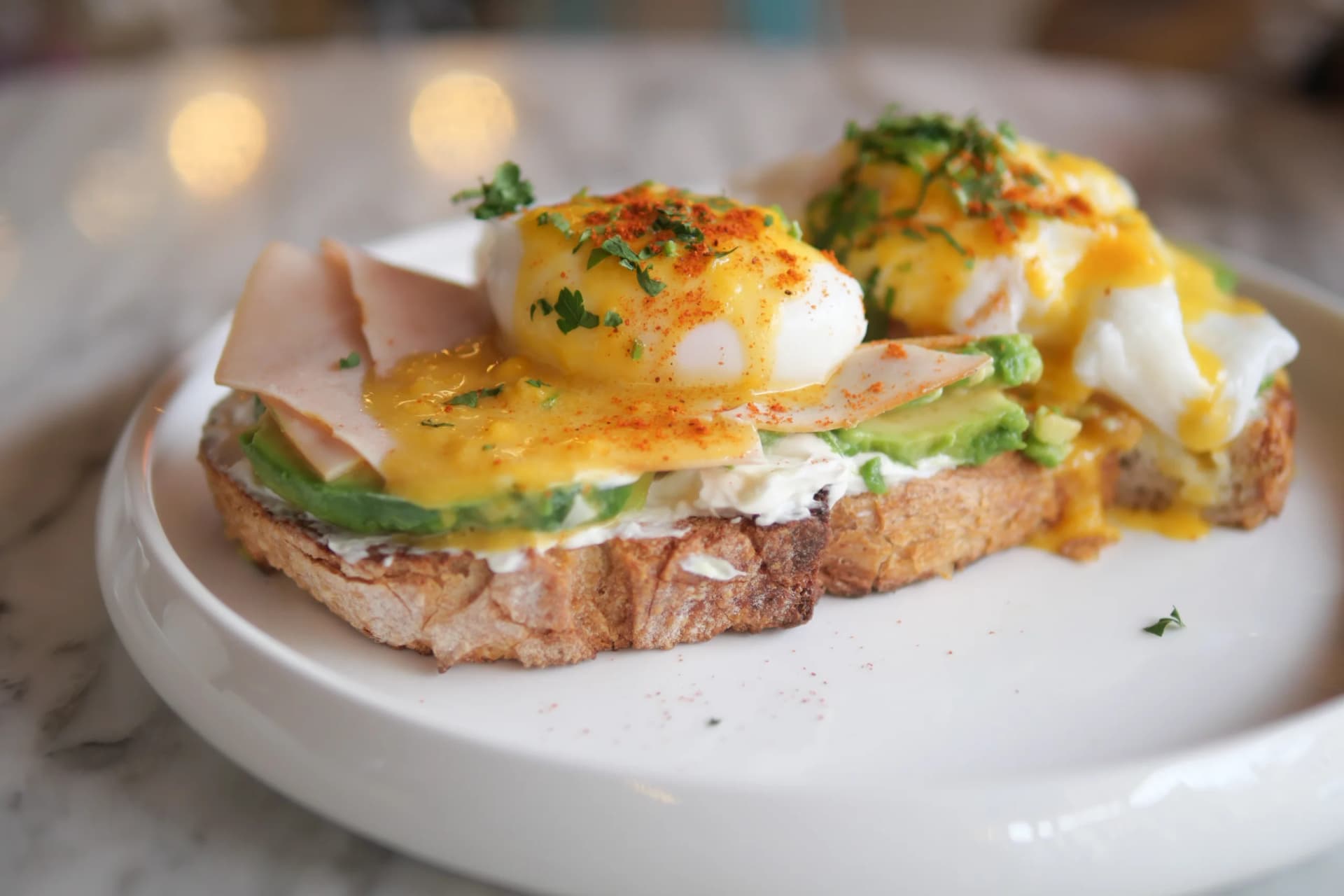 Eggs Benedict with poached eggs, hollandaise sauce, avocado, and turkey on toasted bread, garnished with herbs.