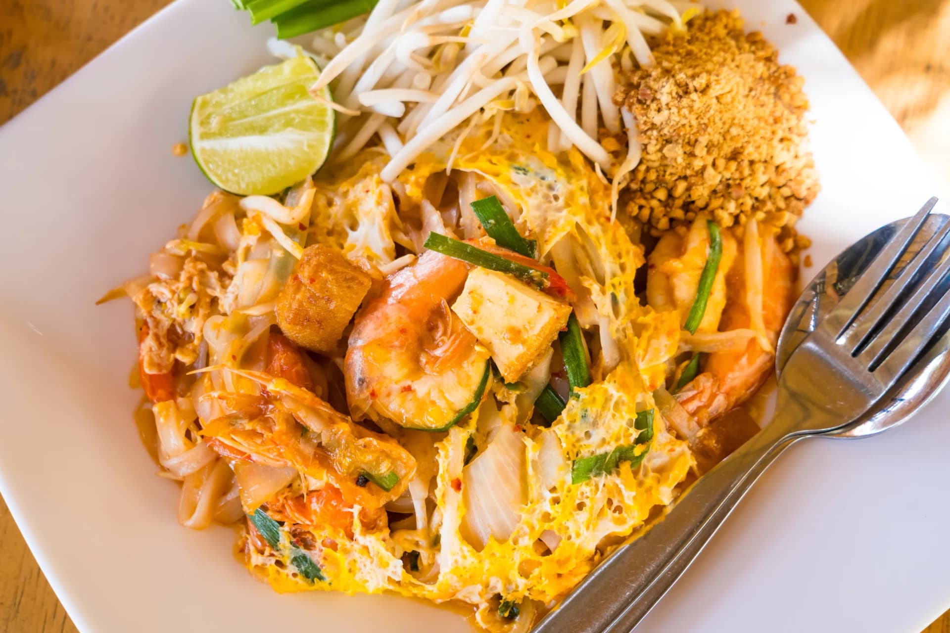 Pad Thai with shrimp, tofu, bean sprouts, lime, and crushed peanuts on a white plate with a fork and spoon.